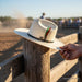 White cowboy hat with a band and plume on a wooden post, with a blurred background of people and a dirt field.