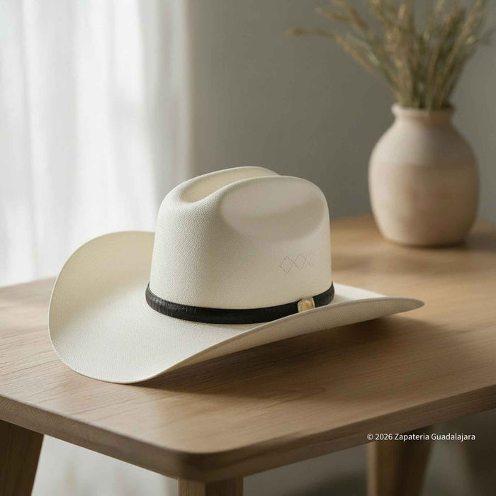 A cream-colored felt cowboy hat with a black band and small gold accent, resting on a light wooden table in a softly lit interior setting.