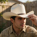 A man wearing a light beige cowboy hat with a dark brown band and a tan button-up shirt, holding the brim of the hat with his hand in an outdoor setting with a wooden fence in the background.