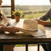 A light beige woven straw cowboy hat with a decorative lattice pattern on the crown and a dark band, resting on a rustic wooden table. A person's hands are adjusting the brim, with a woman and a man seated at the table in a warm, sunlit setting with a blurred outdoor landscape in the background.