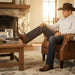 Man in cowboy hat and boots sitting in a leather chair with a fireplace and books in the background