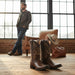 Brown cowboy boots on a wooden floor with a man and chair in the background.