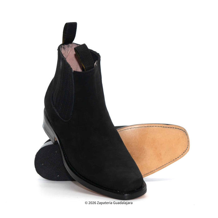 A black suede Chelsea boot shown from the side, with its sole and tan insole visible behind it, on a clean white background.