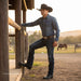 A man wearing a dark blue long-sleeve button-up shirt, dark blue jeans, a brown leather belt with a large silver buckle, and black cowboy boots leans against a wooden post at a ranch, with horses and a fence visible in the background.