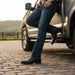 A man wearing dark blue jeans and black leather chukka boots leans against the side of a silver pickup truck on a cobblestone road during golden hour, with a blurred rural landscape in the background.