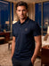 Man wearing a navy polo shirt with a logo in a room with large windows and cityscape view.