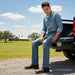 A man wearing a light blue denim shirt and matching blue jeans sits in the bed of a pickup truck outdoors on a gravel road, with a grassy field, trees, and a barn in the background under a blue sky.