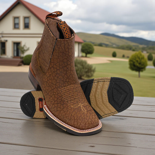 Brown leather boots with black soles on a wooden surface, with a scenic background of a house and greenery.