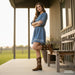 Woman in a blue dress and brown boots standing on a wooden porch with a bench.