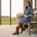 Woman in a blue dress and brown boots sitting on a wooden bench with a scenic background.