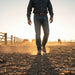 A man wearing a dark button-up shirt, blue jeans, and brown boots walks through a dusty ranch paddock at sunset, with fences and horses visible in the background.