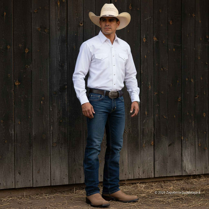 A man wearing a white long-sleeve western shirt, blue jeans, a beige cowboy hat, and brown boots stands against a dark weathered wooden fence.