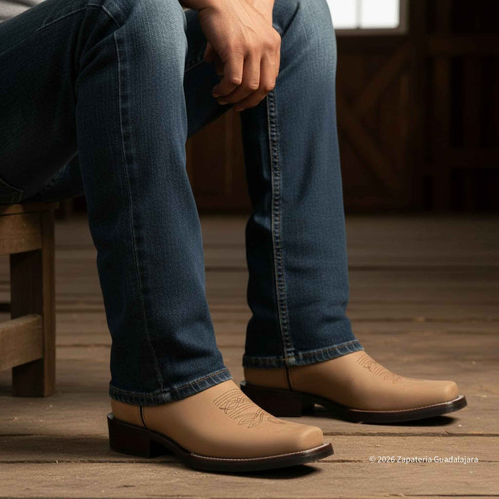 A person seated on a wooden stool wearing blue jeans and tan leather cowboy boots with dark soles, photographed in a rustic indoor setting with wood flooring.
