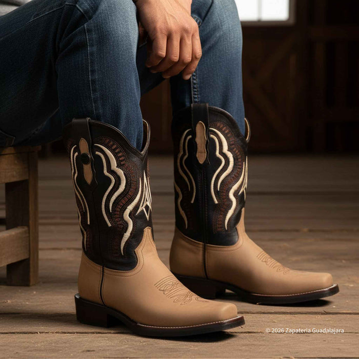 A person seated on a wooden stool wearing tan and black western cowboy boots with white embroidered detailing, paired with blue jeans in a rustic indoor setting.