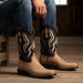 A person seated on a wooden stool wearing tan and black western cowboy boots with white embroidered detailing, paired with blue jeans in a rustic indoor setting.