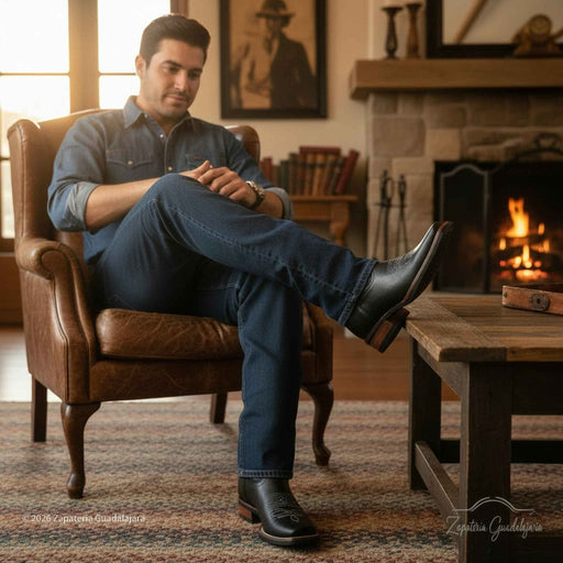 Man sitting in a brown leather chair by a fireplace wearing blue jeans and black boots.