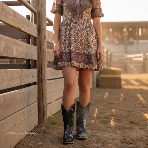Person wearing a floral dress and cowboy boots standing near a wooden fence.