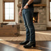 Person wearing blue jeans and black shoes standing in a room with a stone fireplace.
