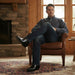 Man sitting in a brown leather chair in a cozy room with stone fireplace and large window.
