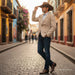 A man wearing a beige blazer, blue jeans, brown boots, and a brown cowboy hat walks down a cobblestone street lined with colorful buildings.