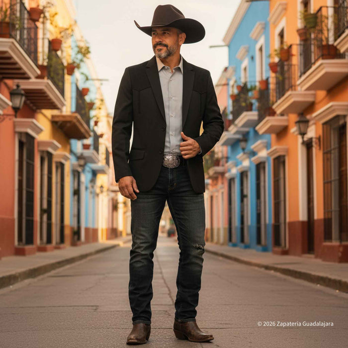 A man wearing a black cowboy hat, black blazer, light blue dress shirt, dark jeans, and brown boots stands on a city street lined with colorful colonial-style buildings.