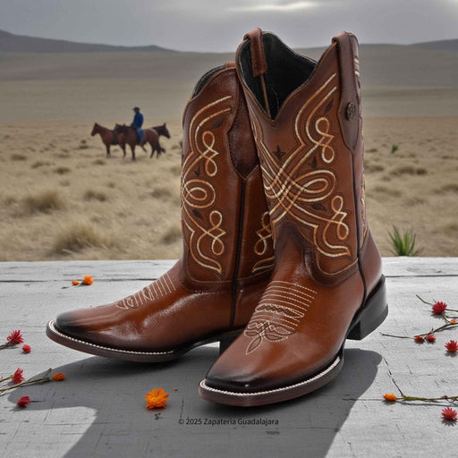 Brown cowboy boots with intricate white stitching on a wooden surface with a desert landscape in the background.