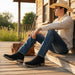 Person sitting on a wooden porch with a guitar, wearing a cowboy hat and boots.