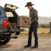 Man in cowboy hat loading a wooden crate into the back of a pickup truck in a rural setting.