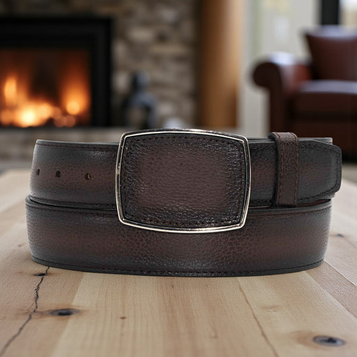 Brown leather belt with a silver buckle on a wooden surface with a fireplace in the background