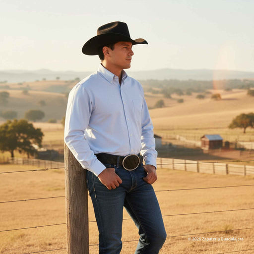 A man in a black cowboy hat, light blue button-down shirt, and dark blue jeans leans against a wooden post in a sunny, rolling pasture landscape.