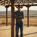 A man wearing a brown cowboy hat, a long-sleeve button-up shirt with a blue and brown paisley pattern, and blue jeans stands with arms crossed under a wooden pergola in an outdoor desert landscape.