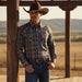 A man wearing a brown cowboy hat, a long-sleeve button-up shirt with a blue and beige paisley pattern, and blue jeans, leaning against a wooden post in an outdoor ranch setting.