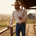 A man wearing a western-style long-sleeve button-up shirt with a beige and brown geometric pattern, blue jeans, and a cowboy hat stands on a wooden porch at sunset with a rural landscape in the background.