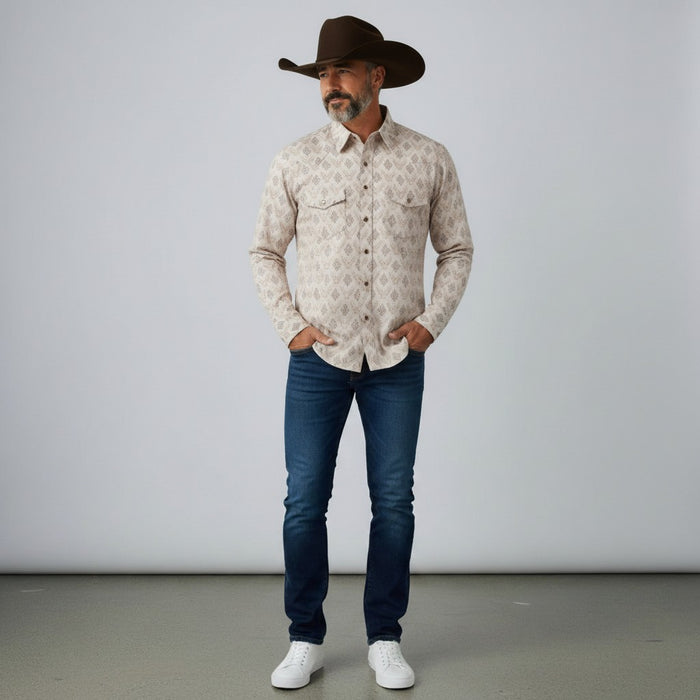 A man wearing a long-sleeve western-style button-up shirt with a beige and brown geometric pattern, dark brown cowboy hat, blue jeans, and white sneakers, standing against a light gray studio background.
