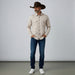 A man wearing a long-sleeve western-style button-up shirt with a beige and brown geometric pattern, dark brown cowboy hat, blue jeans, and white sneakers, standing against a light gray studio background.