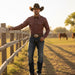 A man wearing a brown cowboy hat, a maroon patterned western shirt, blue jeans, and a large silver buckle belt stands in a sunny field next to a wooden fence, with horses grazing in the background.