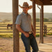 A man wearing a short-sleeve button-up western shirt, dark blue jeans, and a brown cowboy hat leans against a wooden post on a covered porch at sunset, with a horse visible in the background field.