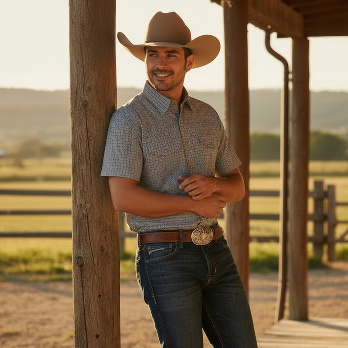 A smiling man wearing a brown cowboy hat, a short-sleeve button-up shirt with a subtle blue and white pattern, and dark blue jeans stands under a wooden porch in a rural outdoor setting.