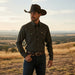A man wearing a dark brown cowboy hat, a patterned long-sleeve western shirt, dark jeans, and a brown belt stands outdoors in a dry grassy field at sunset.