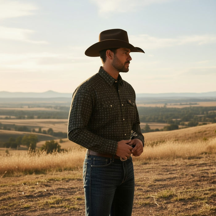 A man wearing a dark brown cowboy hat, a dark patterned long-sleeve button-up shirt, and blue jeans stands on a grassy hill overlooking a vast, rolling landscape under a warm sky.