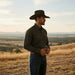 A man wearing a dark brown cowboy hat, a dark patterned long-sleeve button-up shirt, and blue jeans stands on a grassy hill overlooking a vast, rolling landscape under a warm sky.