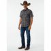 A man modeling a short-sleeve button-down shirt with a dark navy and brown geometric pattern, paired with blue jeans, brown cowboy boots, and a brown cowboy hat against a white studio background.