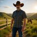 A man wearing a brown cowboy hat and a dark blue patterned short-sleeve button-up shirt stands outdoors in a sunny rural landscape with hills, a fence, and a horse in the background.