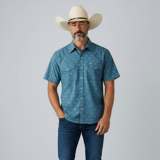 A man wearing a blue short-sleeve button-up shirt with a subtle white pattern, dark blue jeans, white sneakers, and a cream cowboy hat, standing against a light gray studio background.
