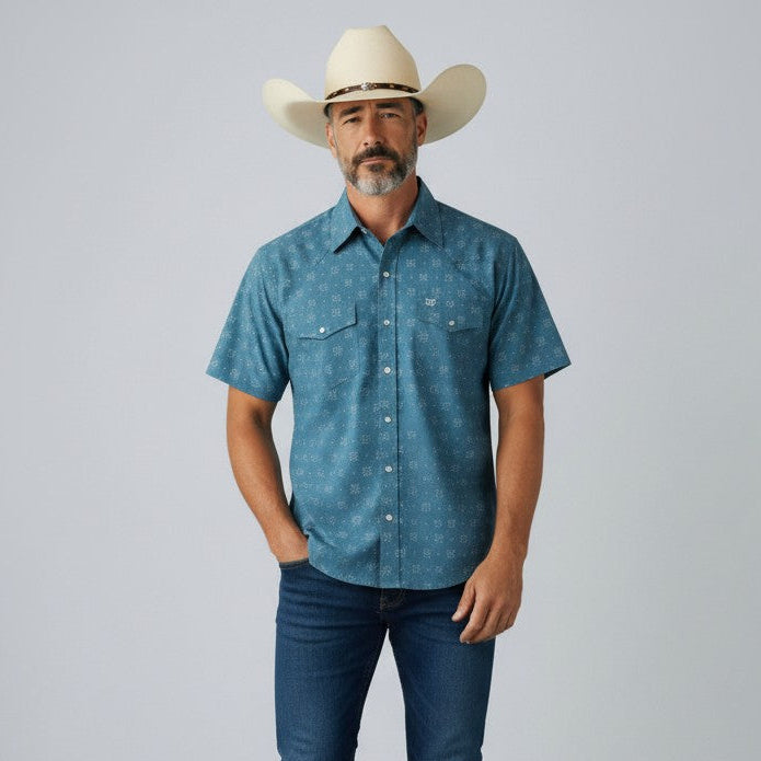 A man wearing a blue short-sleeve button-up shirt with a subtle white pattern, dark blue jeans, white sneakers, and a cream cowboy hat, standing against a light gray studio background.