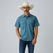 A man wearing a blue short-sleeve button-up shirt with a subtle white pattern, dark blue jeans, white sneakers, and a cream cowboy hat, standing against a light gray studio background.