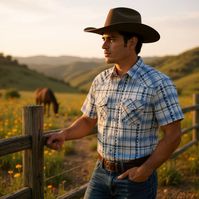 A man wearing a brown cowboy hat, a blue and white plaid short-sleeve button-up shirt, and jeans stands by a wooden fence in a sunny grassy field with hills in the background.