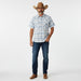 A man modeling a short-sleeve western plaid button-down shirt, dark blue jeans, a brown cowboy hat, and brown boots against a plain white studio background.