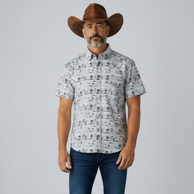 A man wearing a brown cowboy hat, a light gray short-sleeve button-up shirt with a black graphic print, dark blue jeans, and white sneakers, standing against a plain light gray studio background.
