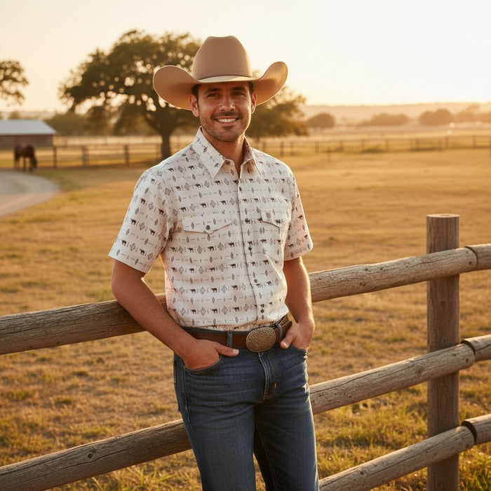 A man wearing a cowboy hat, a short-sleeve button-up shirt with a small black-on-cream pattern, and blue jeans stands outdoors in a rural setting with a wooden fence and field behind him.